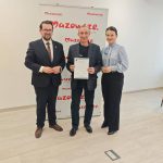 654384665_951498423892346_7873659734318282808_n Trzy osoby, dwie w ciemnych garniturach i jedna w jasnej bluzce, stoją przed ścianką z logo Mazowsze. Jedna osoba trzyma certyfikat. Drewniana podłoga i białe ściany tworzą jasne tło.