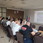 654399095_951496217225900_7666776180077796268_n Na zdjęciu znajduje się grupa osób siedząca przy stole konferencyjnym. Tło stanowi jasna sala konferencyjna z flagami i projektorem. Na ekranie widoczna jest prezentacja o konwencie sołtysów.