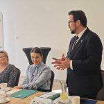 654412620_951497207225801_7535788588041145266_n Na zdjęciu widać mężczyznę w garniturze przemawiającego do grupy osób przy stole. Tło jest jasne z oznakowaniem Gmina Dobre. Na stole są dokumenty i filiżanki z kawą.