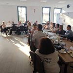 654443743_951496267225895_6993051627895474732_n Na zdjęciu widoczna jest grupa ludzi siedzących przy stołach w jasnym pomieszczeniu. Ściany są białe, z dużym krzyżem i promami Mazowsze. Na stołach leżą notatki i filiżanki z kawą.