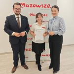 654723594_951497940559061_3666710933464926840_n Trzy osoby stoją przed białym tłem z napisem Mazowsze. Środkowa osoba trzyma dokument. Podłoga jest drewniana.