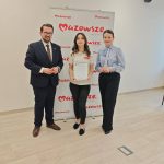 654953617_951498283892360_8907272244671320846_n Trzy osoby stoją przed banerem Mazowsze w jasnym pokoju. Środkowa osoba trzyma certyfikat. Podłoga jest drewniana.