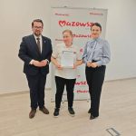 655142092_951498680558987_161234404562069302_n Trzy osoby stoją na tle baneru Mazowsze. Środkowa osoba trzyma certyfikat. Tło jest jasne, podłoga drewniana.