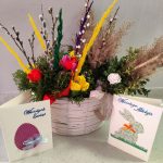 Koszyk z wielkanocną dekoracją z kolorowymi kwiatami i gałązkami na jasnym tle. Obok stoją dwie kartki świąteczne z napisami: „Wesołych Świąt” i „Wesołego Alleluja”. Dekoracje w żywych kolorach podkre