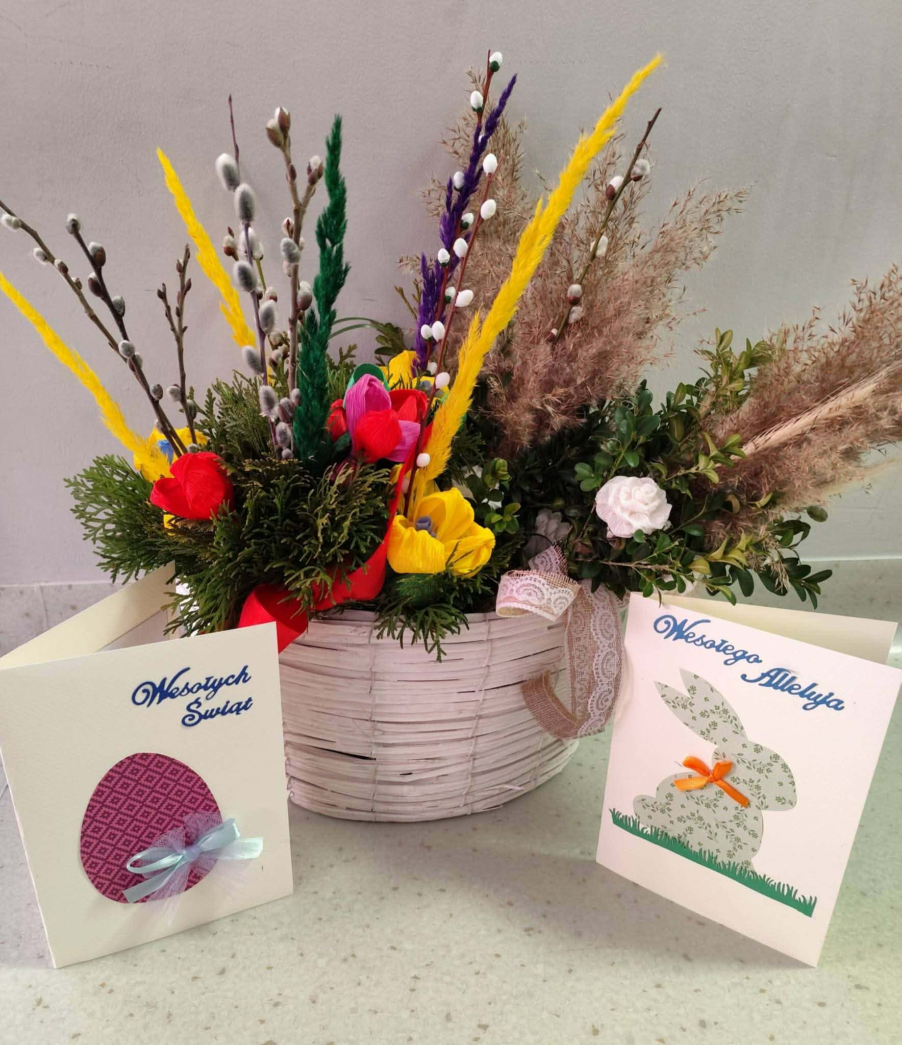 Koszyk z wielkanocną dekoracją z kolorowymi kwiatami i gałązkami na jasnym tle. Obok stoją dwie kartki świąteczne z napisami: „Wesołych Świąt” i „Wesołego Alleluja”. Dekoracje w żywych kolorach podkre
