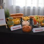 DSC_0318 Na obrazie znajduje się stół z przekąskami: warzywa, dipy, nachosy. Dominuje ciemne tło, jasne zasłony z tyłu. Logo Mazowsze na widocznym znaku.