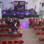 DSC_0323 Scena udekorowana wielobarwnymi kwiatami z instrumentami muzycznymi. Przed sceną czerwone krzesła z widownią. Jasne, przestronne wnętrze z zasłonami.