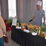 DSC_0328 Na zdjęciu widać kucharza przy stole z jedzeniem. Na stole znajdują się warzywa, oliwa i zioła. Tło stanowią szare zasłony, a obok stoją czerwone krzesła.