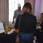 DSC_0329 Grupa ludzi stoi przy stole z jedzeniem na czarnym obrusie. Na stole są kolorowe naczynia oraz bukiet różowych tulipanów. W tle znajduje się kucharz w białym ubraniu.