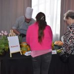 DSC_0333 Na zdjęciu jest kucharz w białej czapce, przygotowujący potrawy przy stole z ziołami i tulipanami. Obok dwie kobiety w jasnych strojach oglądają składniki. W tle szare zasłony i białe światło.