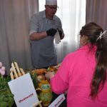 DSC_0334 Kucharz w czapce i fartuchu demonstruje przygotowanie potraw. Kobieta w różowej bluzie obserwuje. Na stole są tulipany i zioła.