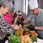 DSC_0335 Na zdjęciu szef kuchni w białej czapce serwuje potrawy. Przed nim stół z pieczywem, warzywami i ziołami. Tło to jasne zasłony i fioletowe światła.