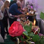 DSC_0339 Czerwona róża na pierwszym planie, z boku grupa ludzi przy stole z jedzeniem. W tle widoczne są świeczki i dekoracje.