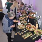 DSC_0341 Stół z różnorodnymi potrawami, kwiaty w wazonach na tle jasnej ściany. Ludzie w kolejce, kolorowe dekoracje. Sala bankietowa w jasnym świetle.