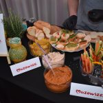 DSC_0344 Na stole widać przekąski: kanapki, warzywa, dipy i pieczywo. Tło jest jasne, ozdobione roślinami. Logo Mazowsze na kartkach.