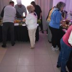 DSC_0346 Grupa ludzi wokół stołu z jedzeniem w jasnym pokoju. Stół pokryty czarnym obrusem, z kolorowymi potrawami. Tło z zasłonami i fioletowym światłem.