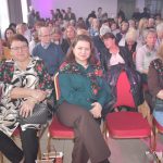 DSC_0350 Na zdjęciu widać grupę osób siedzących na czerwonych krzesłach. Tło jest jasne z zasłonami i fioletowym światłem. Kilka osób nosi kwieciste chusty.