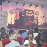 DSC_0354 Na scenie mężczyzna w garniturze przemawia, otoczony różowym i fiołkowym światłem. W tle instrumenty muzyczne, napisy i dekoracje. Publiczność siedzi na czerwonych krzesłach, po lewej stronie biały baner z czerwonym napisem Mazowsze.