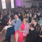 DSC_0358 Sala pełna ludzi siedzących na czerwonych krzesłach, klaszczących. Tło jest jasne, z zasłonami i subtelnym fioletowym oświetleniem. W przodzie osoby w kolorowych strojach.