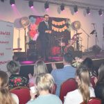 DSC_0359 Mężczyzna w garniturze przemawia na scenie z instrumentami muzycznymi. Tło udekorowane kolorowymi kwiatami i napisem. Widzowie siedzą na czerwonych krzesłach.