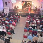 DSC_0362 Na zdjęciu widoczna jest sala z koncertem, pełna osób siedzących na czerwonych krzesłach. Na scenie występujący zespół, tło ozdobione kolorowymi kwiatami i napisem. Pomieszczenie oświetlone ciepłym światłem, w tle biał