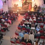 DSC_0363 Widok z góry na koncert w jasnej sali. Publiczność siedzi na czerwonych krzesłach. Na scenie zespół i wokalistka w czerwonej sukni.