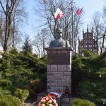 Popiersie na cokole z brązowego kamienia, otoczone zielonymi krzewami. Widoczne polskie flagi i wiosenne dekoracje. Tło to nagie drzewa i fragment budynku z cegły.