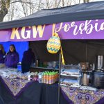 Stragan z fioletowym napisem, oferujący lokalne produkty spożywcze. Na pierwszym planie ciasta i napoje, ozdobiony wzorzystymi obrusami. Tło wypełniają drzewa i niebieskie płachty.