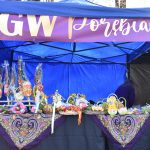 Niebieski namiot wystawowy z plecionymi koszami i ozdobami na stole. Fioletowy szyld z napisem na górze. Tło to ciemnoniebieska plandeka.