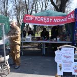 Mężczyzna w stroju strażaka gotuje zupę w czarnym garnku nad ogniem. W tle zielony namiot i osoby przy stole. Kolorowy stragan z rękodziełem z boku.