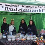 Cztery kobiety w ciemnych strojach stoją przed zielonym namiotem. Na stole leżą zapakowane ciasta i ozdoby wielkanocne. Baner nad nimi głosi Rudzienianki.