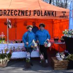 Na zdjęciu widoczny jest pomarańczowy namiot z napisem i stoisko z kwiatami. Trzy osoby w niebieskich kurtkach i wiankach stoją z bukietami. Tło stanowi zalesiony teren.