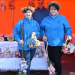 Trzy osoby w niebieskich kurtkach stoją przed pomarańczowym namiotem, trzymając ozdobne miotły. W tle stół z kwiatowymi dekoracjami. Na głowach mają kwiatowe wianki.