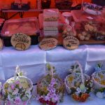 Kolorowe stoisko z wypiekami w plastikowych pudełkach na białym obrusie. Drewniane znaki z nazwami produktów. U dołu koszyki z kwiatami.