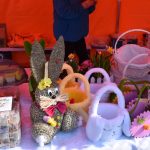 Na zdjęciu stół z ozdobami świątecznymi, w tym wiklinowy zając z kokardą i koszyki. Tło pomarańczowe, na stole różowe i białe dekoracje. Obok są ciasta w plastikowych opakowaniach i kolorowe kwiaty.