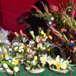 Stół z kolorowymi dekoracjami wielkanocnymi. W tle czerwony materiał i ciemno ubrana postać. Na stole ozdoby z kwiatami i kurczakami.