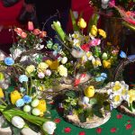 Kolorowe kompozycje wielkanocne na zielonym obrusie. Żółte kurczaczki, jajka i kwiaty tulipanów dominują w aranżacji. Tło w czerwieni dodaje świątecznego klimatu.