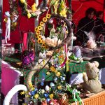 Ręcznie robione wieńce z kwiatami i wstążkami wiszą na drzewie. Tło w kolorze czerwonym z zielonym stolikiem. Obok jest pleciony kosz z ozdobnym misiem.