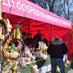 Na zdjęciu widoczny jest jaskraworóżowy namiot targowy z wiejskimi produktami. Stoisko zdobią kolorowe wieńce i kwiaty. W tle drzewa, a ludzie oglądają wystawione produkty.