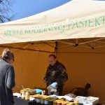 Stanowisko z miodem, namiot z napisem, mężczyzna w rękawiczkach sprzedaje różnokolorowe słoiki.