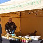 Mężczyzna w kamuflażu sprzedaje miody w żółtym namiocie. Przed nim słoiki z miodem na czarnym stole. Napis: MIÓD Z RODZINNEJ PASIEKI.