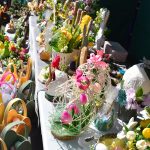 Kolorowe rękodzieła na targu wielkanocnym zdobią stół. Dominują ozdoby z kwiatami, jajkami i zajączkami. Tło zielone, otoczone wiosennymi dekoracjami.