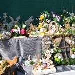 Stół pełen wielkanocnych ozdób z zajączkami i kwiatami. Dominuje zielony, różowy i kremowy kolor. Tło ciemnozielone.