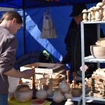 Dwie osoby na targu oglądają drewniane produkty. Stoisko jest pod niebieskim namiotem. Drewniane naczynia są ustawione na półkach.