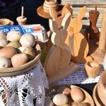 Drewniane jajka w misie na białej koronkowej serwecie. W tle drewniane ozdoby w kształcie królików. Jasne kolory drewna dominują.