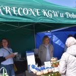 Na zdjęciu widoczny jest zielony namiot z napisem oraz stoisko z domowymi wypiekami. Dwie kobiety obsługują klientów. Namiot otoczony jest innymi w kolorze niebieskim.