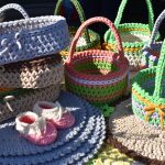 Kolorowe, ręcznie robione koszyki z włóczki i różowe buciki na tle granatowej powierzchni. Koszyki mają różne kolory, w tym zielony, różowy i beżowy. Po prawej stronie widać również kilka okrągłych podkładek