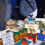 Stoisko z przetworami i rękodziełem. Słoiki z kolorowymi zakrętkami, tace, dekoracje. Podkładki i kosze na tle ciemnej tkaniny.