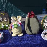 Na zdjęciu widać ręcznie robione ozdoby wielkanocne z włóczki w pastelowych barwach, na ciemnoniebieskim tle. W tle znajduje się zielona ściana namiotu, co podkreśla rękodzieło. Elementy dekoracyjne przedstawiają motywy zwier