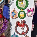 Na zdjęciu widoczne są kolorowe, ozdobne wianuszki wiszące na białej desce. Wianuszki mają różne motywy, pastelowe odcienie i dodatki jak kokardki, kwiaty czy figurki zwierząt. W tle jasno oświetlone tło